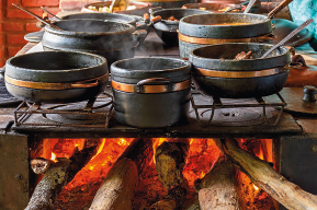 Imagem: Fotografia. Destaque de panelas de ferro no fogão a lenha aceso. Fim da imagem.