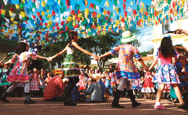 Imagem: Fotografia. Destaque de um grupo de crianças que dançam quadrilha em uma área externa enfeitada com muitas bandeirolas coloridas presas formando varais bem longos. As meninas estão de pé e forma um círculo de mãos dadas. Elas usam vestidos coloridos, meias, sapatos e chapéu ou fitas na cabeça. Os meninos estão sentados ao centro da roda. Eles usam chapéu de palha, camisa xadrez, calça e tênis.  Fim da imagem.