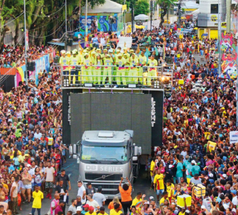 Imagem: Fotografia. Destaque de uma via tomada por uma multidão ao redor de um trio elétrico sobre o qual há um grupo de pessoas que usam um mesmo uniforme verde e branco. Fim da imagem.