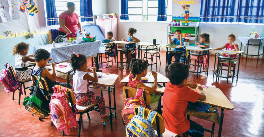 Imagem: Fotografia. Destaque do interior de uma sala de aula com crianças sentadas em carteiras posicionadas em formato semicircular, incluído o espaço da professora, que está de pé. As mochilas estão penduradas nas cadeiras. Há colagem nas paredes e cortinas nas janelas.  Fim da imagem.