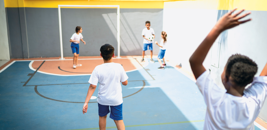 Imagem: Fotografia. Em uma quadra descoberta, estão cinco crianças uniformizadas. Elas estão de pé e uma segura uma bola nas mãos. Fim da imagem.