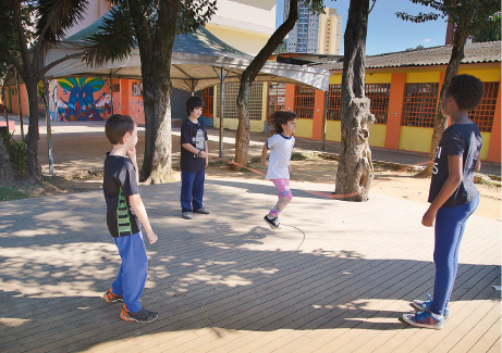 Imagem: Fotografia. Em um espaço aberto e sombreado por árvores, estão quatro crianças. Duas batem corda, uma pula e outra observa.  Fim da imagem.