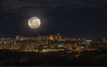 Imagem: Fotografia em plano aberto. Paisagem urbana composta por muitos prédios com luminosidade. No céu límpido escuro, a lua cheia.  Fim da imagem.
