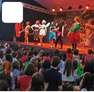 Imagem: Fotografia. Um grupo de pessoas de características diversas, adultos e crianças, observa diferentes personagens fantasiados em um palco iluminado e decorado. Fim da imagem.