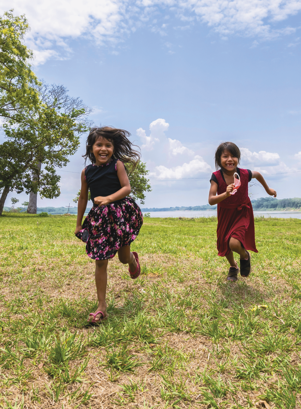 Imagem: Fotografia. Duas meninas de aproximadamente seis anos sorriem enquanto correm em uma área gramada com árvores ao redor e um rio ao fundo. Elas têm pele parda e cabelo liso e preto. Uma está com vestido preto regata com flores rosas e chinelo e a outra está com longo vestido vermelho de manga curta e tênis e segura uma máscara. Fim da imagem.