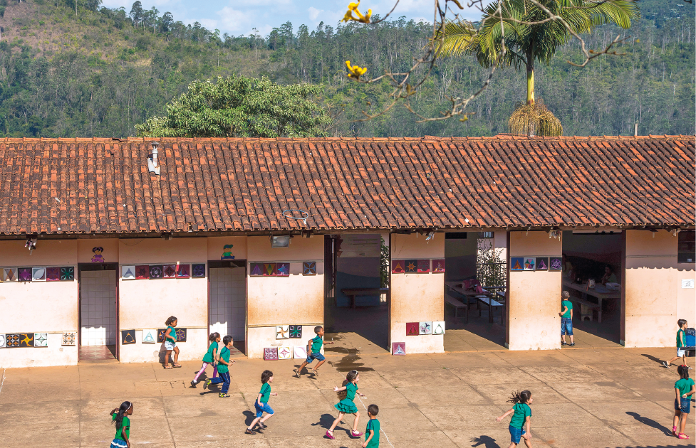 Imagem: Fotografia em plano aberto. No pátio da escola, crianças uniformizadas correm e caminham na mesma direção. Elas estão diante das paredes dos banheiros, identificados com a figura de um menino e de uma menina, e ao lado estão as portas das salas de aula e refeitório. O telhado é de cerâmica e há um bambolê próximo à borda. Ao fundo, muitas árvores. Fim da imagem.