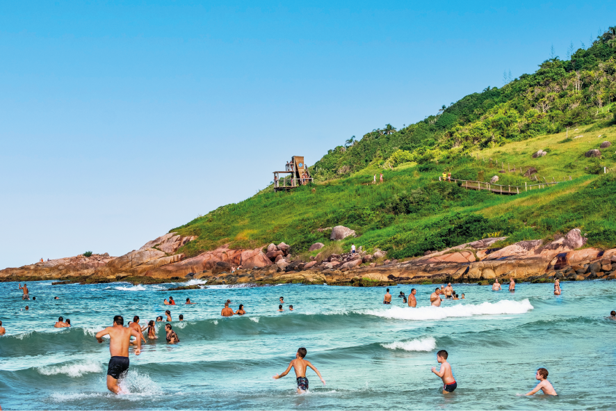 Imagem: Fotografia. Muitas pessoas, homens, mulheres e crianças, divertem-se na água do mar em uma praia rodeada por pedras e uma serra gramada na qual passa uma pequena ponte de madeira. No alto, há uma pequena construção de madeira de onde pessoas observam a água. O céu está azul.  Fim da imagem.
