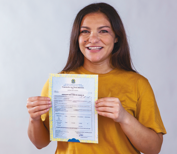 Imagem: Fotografia. Retrato de uma mulher de pele clara, cabelo castanho-escuro liso e longo, solto. Ela sorri e segura com as duas mãos uma certidão de nascimento à frente do corpo. Fim da imagem.