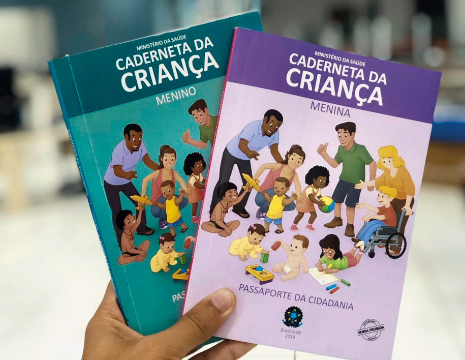 Imagem: Fotografia. Destaque da mão de uma pessoa que segura dois materiais impressos identificados como “Caderneta da criança”, um é roxo e apresenta a inscrição “Menina” e o outro é azul, e apresenta a inscrição “Menino”. Em ambos, a capa traz diversas crianças, de diferentes características, exercendo diferentes atividades.  Fim da imagem.