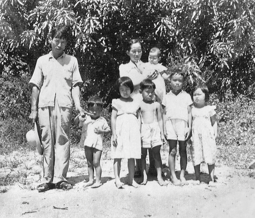 Imagem: Fotografia em preto e branco. Um homem segura um chapéu em uma das mãos e do seu lado esquerda está de mãos dadas com uma criança pequena. Ao lado deles, estão uma mulher com uma criança de colo e à sua frente mais quatro crianças maiores, dois meninos e duas meninas. Eles têm olhos ligeiramente puxados e cabelo escuro e liso. Eles estão em uma área aberta com solo terroso e à frente de árvores. Fim da imagem.