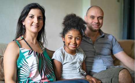 Imagem: Fotografia. Uma mulher branca de cabelo liso preto, uma menina negra de cabelo preto crespo e um homem branco calvo estão sentados em um sofá e sorriem. Fim da imagem.
