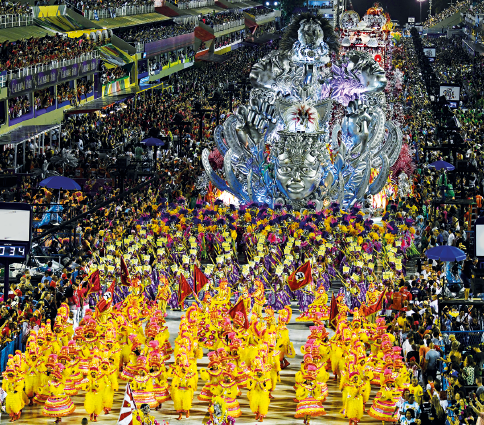 Imagem: Fotografia em plano aberto. Destaque da avenida do sambódromo na qual passam alas com passistas e carros alegóricos com muitas cores. Próximo da via, há camarotes com pessoas de pé e mais ao fundo estão as pessoas nas arquibancadas.  Fim da imagem.