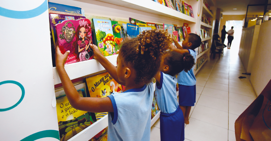 Imagem: Fotografia. Três crianças uniformizadas escolhem livros em uma extensa prateleira. Ao fundo, um corredor comprido e duas pessoas paradas na porta.  Fim da imagem.