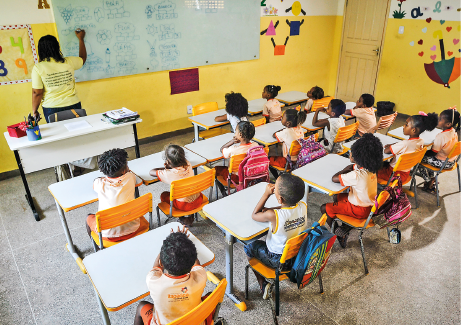 Imagem: Fotografia. Destaque do interior de uma sala de aula na qual as crianças estão sentadas à carteira lado a lado e observam a professora escrever na lousa. Há colagens nas paredes e as mochilas estão penduradas nas cadeiras. Fim da imagem.
