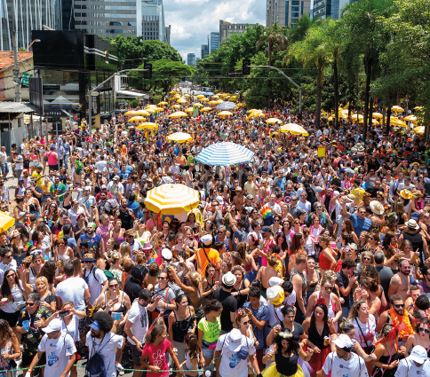 Imagem: Fotografia em plano aberto. Destaque de uma multidão que toma uma ampla via com árvores e prédios ao redor. Entre as pessoas há alguns ambulantes com guarda-sóis.  Fim da imagem.