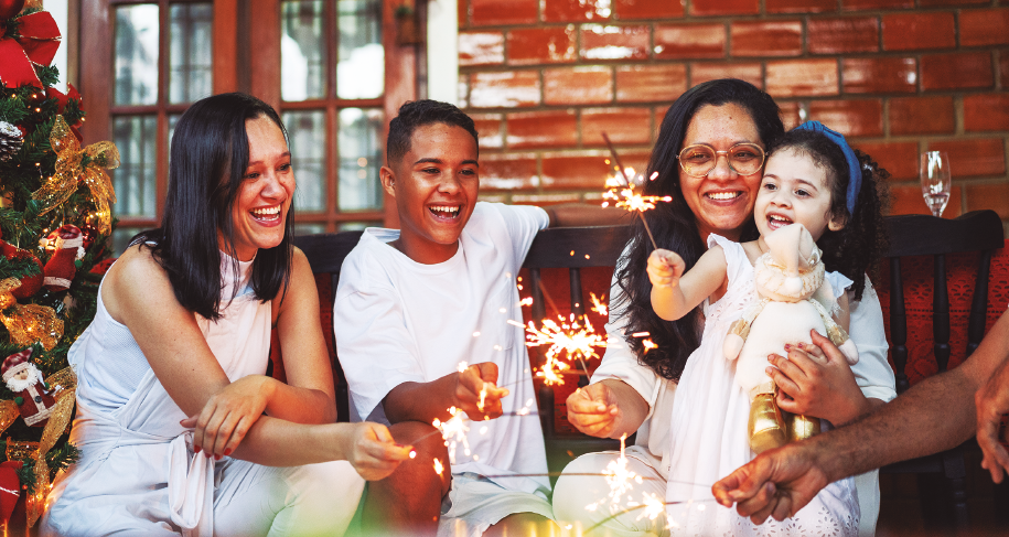Imagem: Fotografia. Em uma sala, ao lado de uma árvore de Natal, duas mulheres adultas e duas crianças vestidas de branco sorriem enquanto seguram finas velas pretas que soltam faíscas douradas.  Fim da imagem.