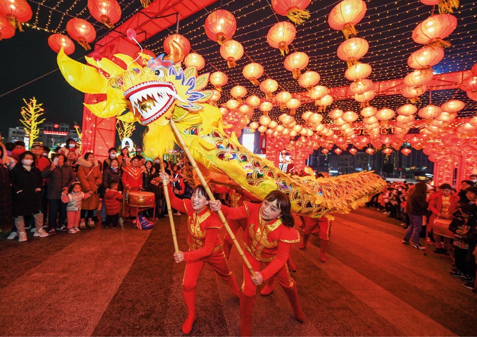 Imagem: Fotografia em plano aberto. Durante à noite, em uma via, ao centro em destaque, duas meninas fantasiadas com roupas vermelhas seguram hastes presas à cabeça de um grande dragão amarelo que apresenta corpo longo e com aspecto escamoso brilhante. Ao longo do corpo, há outras pessoas que vestem roupa vermelha, mas que estão com a cabeça coberta no interior do corpo do animal e apenas os pés de fora.  O espaço por onde passa a montagem apresenta uma estrutura com teto iluminado com muitas luminárias redondas vermelhas. Ao redor, muitas pessoas observam a cena.  Fim da imagem.