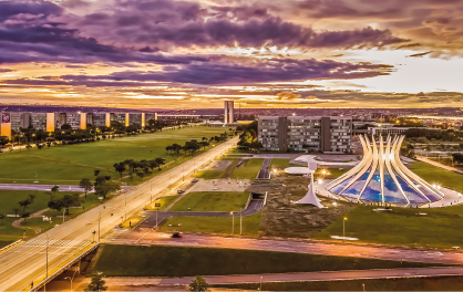 Imagem: Fotografia em plano aberto. Paisagem urbana composta por uma extensa via, área gramada e um prédio circular com arquitetura com colunas diagonais que formam um círculo ao centro. No céu, a luz do sol no horizonte e muitas nuvens. Fim da imagem.
