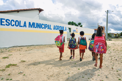 Imagem: Fotografia. Destaque de uma área com chão de terra à frente da Escola Municipal Agrícola e por onde caminham cinco crianças com roupas leves e mochila nas costas. Fim da imagem.