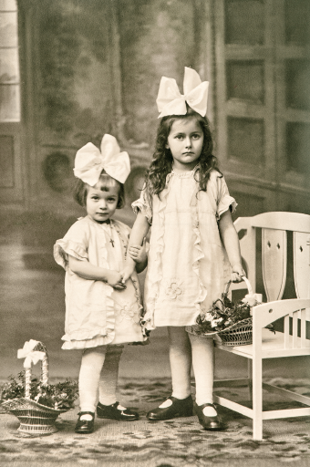 Imagem: Fotografia. Duas meninas posam de mãos dadas para foto com vestidos claros e com grandes laços que enfeitam a cabeça. Elas estão lado a lado. A maior segura um cesto sobre um banco e há outro cesto no chão ao lado da menor.  Fim da imagem.
