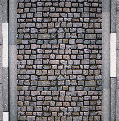 Imagem: Fotografia. Vista superior do destaque de uma via com paralelepípedo.  Fim da imagem.