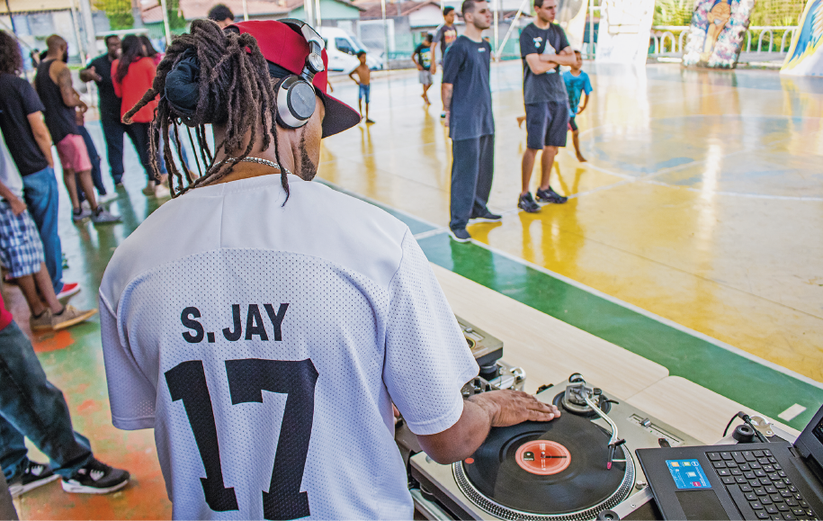 Imagem: Fotografia. Em primeiro plano, destaque de um rapaz de costas que manipula um vinil num toca-discos ao lado de um notebook. Ele tem dreads, está de boné, usa fone de ouvido e usa uma camiseta com inscrição “S. JAY 17”. Ao fundo, jovens parados conversam em uma quadra poliesportiva.  Fim da imagem.