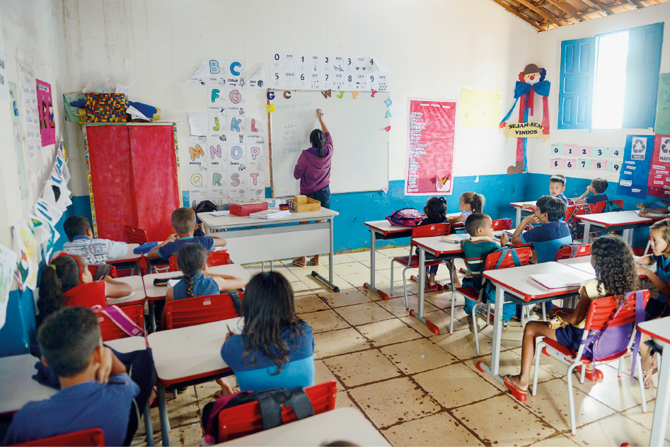 Imagem: Fotografia. Interior de uma sala de aula nas cores branca e azul onde está um grande grupo de crianças uniformizadas sentadas em duplas nas carteiras dispostas enfileiras. Elas estão com estojos e cadernos sobre a mesa, as mochilas estão penduradas nas cadeiras e elas observam a lousa, onde a professora escreve. Na sala ainda há a mesa da professora, com muitos materiais na bancada e um armário com TNT vermelho com caixas dispostos na parte superior. Nas paredes estão colados cartazes, quadros, desenhos, numerais e letras do alfabeto. A telha é de cerâmica e as janelas são de madeira e estão abertas.  Fim da imagem.