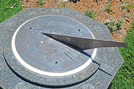 Imagem: Fotografia. Relógio de sol. No chão gramado, uma estrutura redonda que apresenta uma haste fina em relevo e posicionada de forma reta do centro a uma das extremidades da estrutura. Toda a superfície pega sol, com exceção da parte coberta pela espessura da haste, que forma uma linha sombreada sobre o relógio.   Fim da imagem.