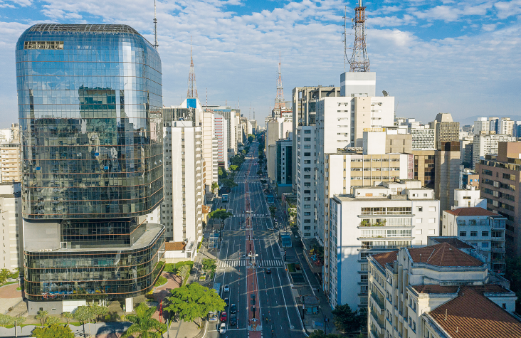 Imagem: Fotografia. Plano aberto de uma extensa via asfaltada que apresenta ciclovia ao centro e é rodeada por altos edifícios, alguns deles com antenas compridas.  Fim da imagem.