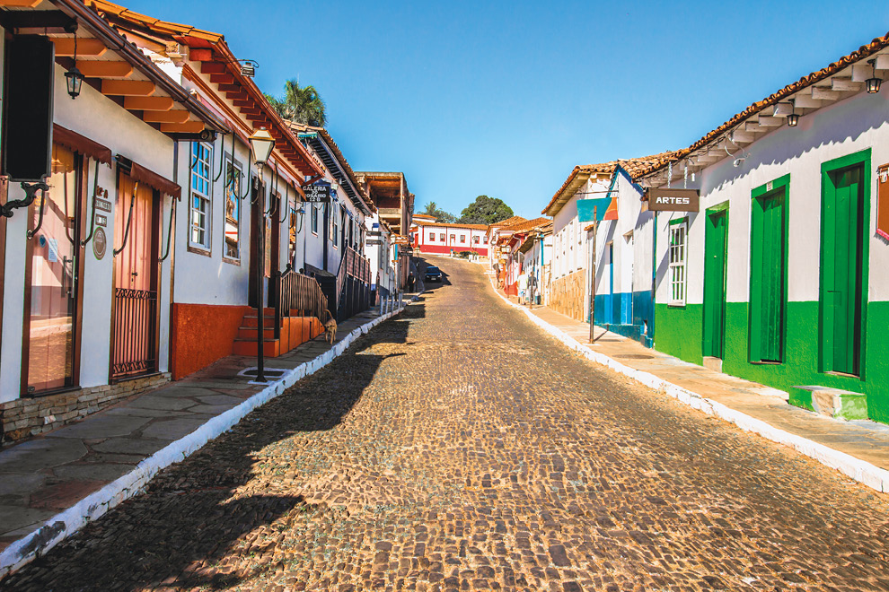 Imagem: Fotografia. Uma estreita via de pedras e pequenas calçadas com postes de iluminação e fachadas de estabelecimentos térreos com portas e janelas de madeira coloridas com acesso direto à rua.  Fim da imagem.