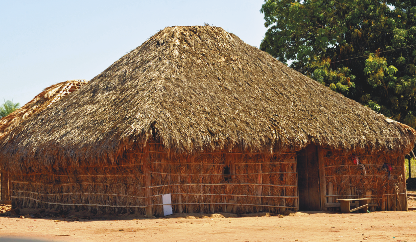 Imagem: Fotografia. Em uma área com chão de terra, uma casa quadrada com paredes firmes de palha e teto também de palha em forma de “V” invertido. Fim da imagem.