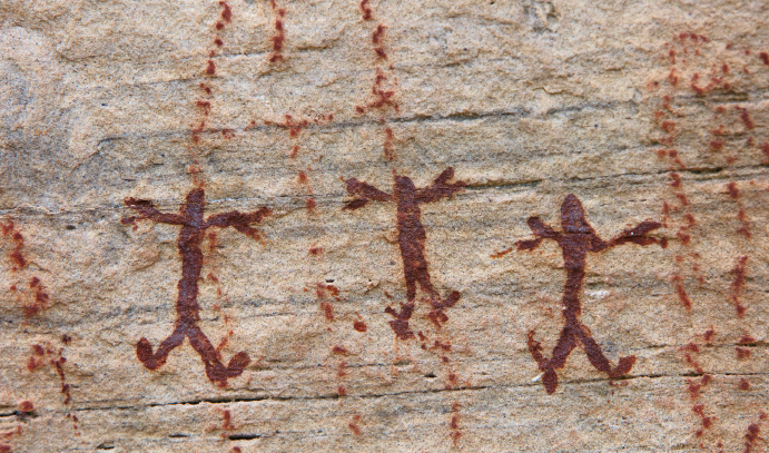 Imagem: Fotografia. Destaque de uma parede rochosa com desenhos de um conjunto de seres com cabeça pequena e corpo alongado e membros logos. Fim da imagem.