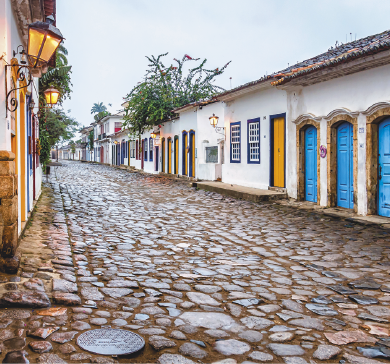 Imagem: Fotografia. Uma rua com pedras e construções térreas com fachadas compostas por portas e janelas de madeira coloridas e com acesso direto à via. Próximo às portas, estão instaladas arandelas. Fim da imagem.