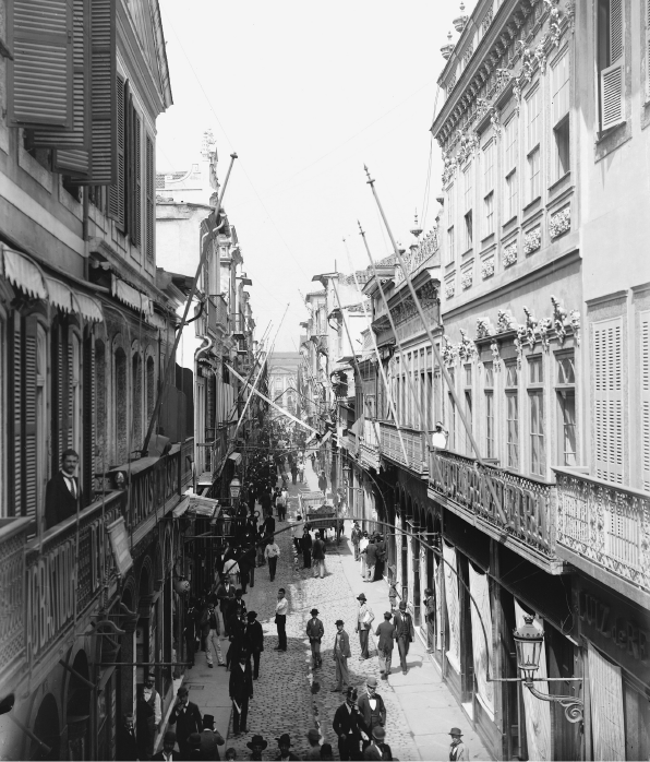 Imagem: Fotografia em preto e branco. Vista de uma estreita rua cercada por edifícios com muitas janelas amplas e com sacadas. Muitas pessoas transitam pela via.  Fim da imagem.