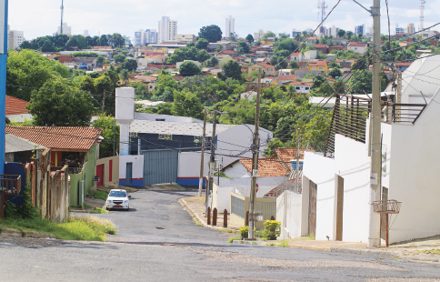 Imagem: Fotografia. Destaque de uma rua pavimentada em uma região de ladeira. No local, há casas e árvores.   Fim da imagem.