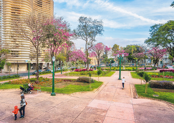 Imagem: Fotografia. Uma praça que apresenta ampla área de passeio cercada por gramado com árvores e postes de iluminação. Poucas pessoas transitam pelo espaço. Ao redor, há um alto edifício.  Fim da imagem.