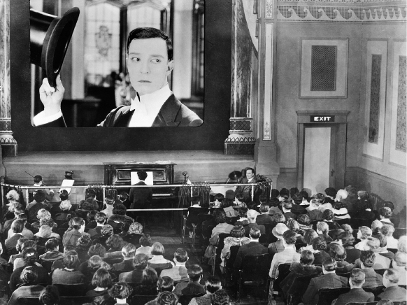Imagem: Fotografia em preto e branco. Espaço interno com muitas pessoas sentadas em cadeiras enfileiras e separadas por um corredor. Elas olham para uma grande tela em piso superior e na qual está a imagem de um homem de terno segurando um chapéu com expressão séria. Entre a plateia e o piso superior, há um rapaz tocando piano e outros instrumentistas. Fim da imagem.