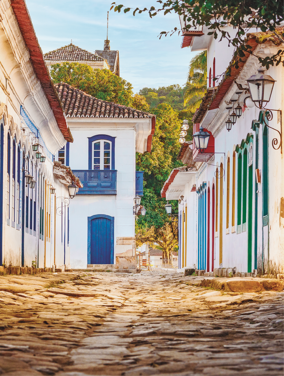 Imagem: Fotografia. Destaque de uma rua estreita calçada com pedras e com construções de fachadas antigas, com portas e janelas de madeira e coloridas com acesso direto à rua e arandelas ao lado das portas. Ao fim da rua, uma construção assobradada com sacada e árvores. Fim da imagem.