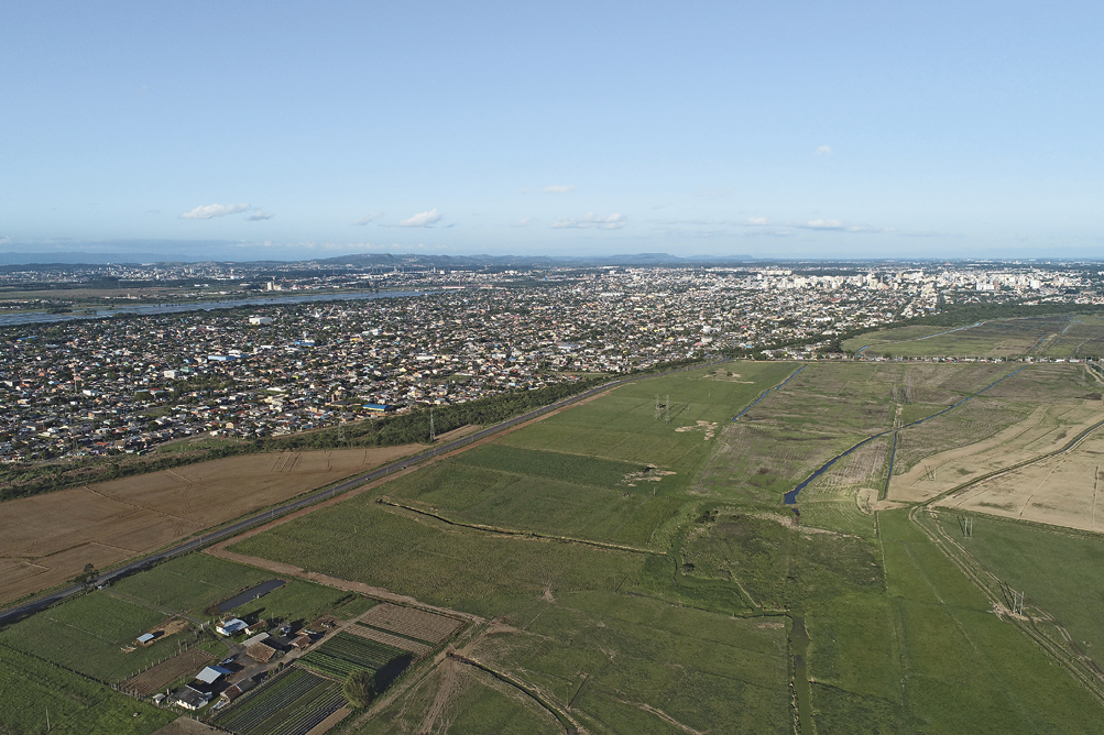 Imagem: Fotografia. Vista aérea de uma extensa área verde com uma estrutura de casas com plantação e um reservatório de água ao redor de uma região descampada e próxima à divisa de uma cidade. No horizonte, morros e o céu.  Fim da imagem.