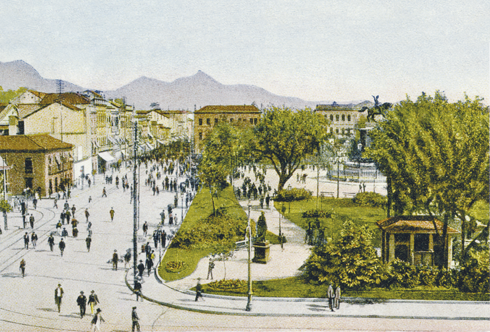 Imagem: Pintura. Plano aberto de uma praça pública arborizada, com coreto, bancos, espaço de passeio e um amplo monumento ao centro. Algumas pessoas transitam no espaço. Do lado esquerdo, uma via sem tráfego é ocupada por pedestres. Há a delimitação da linha férrea do bonde e alguns postes de iluminação. Ao fundo, uma rua estreita com fachadas assobradas e toldos onde o movimento de pessoas é maior. No horizonte, morros.  Fim da imagem.