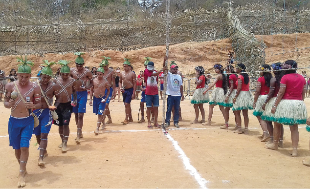 Imagem: Fotografia. Um grupo de pessoas indígenas caminha em fileira formando um semicírculo em um terreno. Do lado esquerdo estão os homens. Eles estão sem camisa, descalços, usam adereços na cabeça, seguram chocalhos, têm faixas pintadas transpassadas na lateral do corpo, usam bermuda e adereços nas pernas. Do lado direito estão as mulheres. Elas usam camiseta de manga curta, saia de palha, estão descalças, tem o braço pintado, usam adereços na cabeça e pernas. Ao centro, estão dois homens de camisa, calça e boné que observam. Ao fundo, uma grade revestida com palha.  Fim da imagem.