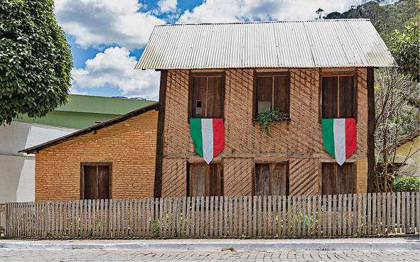 Imagem: Fotografia. Fachada de uma construção assobradada com tijolos aparentes e madeira. As janelas são amplas e verticalizadas e há duas bandeirolas com faixa verde, branca e vermelha penduradas. A casa é rodeada por cerca baixa de madeira e há árvores ao redor.  Fim da imagem.