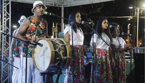 Imagem: Fotografia. Em um palco coberto durante a noite, diante de microfones, um grupo composto por um homem com um tambor e três mulheres no vocal se apresenta. Eles usam roupas com uma composição de parte branca e parte com flores de chita e o homem usa chapéu. Fim da imagem.