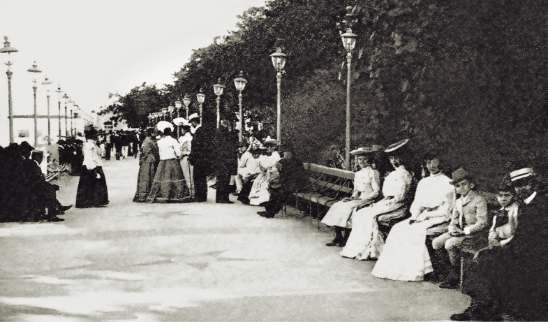 Imagem: Fotografia em preto e branco. Uma área aberta, com largo espaço para passeio e bancos compridos nas laterais ao lado de postes de iluminação. Do lado direito, em destaque, um homem de terno e chapéu posa para foto ao lado de dois meninos, também de terno e chapéu, e três mulheres que usam longo vestido e chapéus grandes.  Ao fundo, outras pessoas conversam em diferentes grupos, algumas de pé e outras sentadas. Fim da imagem.