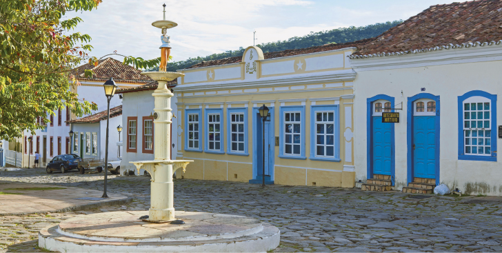 Imagem: Fotografia. Uma rua com paralelepípedos e construções térreas com fachadas compostas por portas e madeiras com acesso direto à rua. Em destaque, há uma fonte ao centro da via.  Fim da imagem.