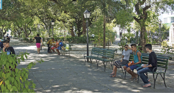 Imagem: Fotografia. Uma menina de saia e camiseta e dois meninos de camiseta, shorts e calça estão sentados no banco de uma praça com largo espaço para passeio, postes de iluminação e muitas árvores. Ao fundo, há outras pessoas também nos bancos. Fim da imagem.