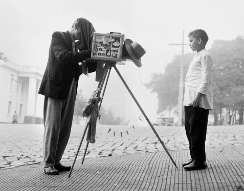 Imagem: Fotografia em preto e branco. Um homem de terno, calça e sapato está de pé diante de uma máquina fotográfica sobre um tripé. Ele está com o rosto coberto por um pano ligado à máquina e diante de um menino que posa de sapato, camisa e calça à sua frente. Um chapéu está pendurado no equipamento  Fim da imagem.