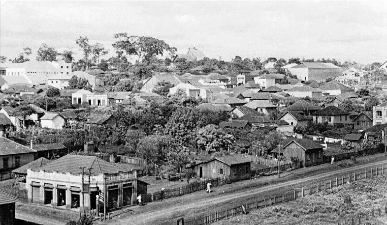 Imagem: Fotografia em preto e branco. Trecho de uma cidade com muitas casas, majoritariamente térreas, entre árvores e uma via de terra ao lado de um terreno com cerca de madeira.  Fim da imagem.