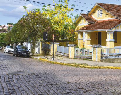 Imagem: Fotografia. Em uma rua com paralelepípedos, destaque de uma casa assobrada de esquina que apresenta uma área com estrutura de duas colunas, telhado em “V” invertido de cerâmica e com muro e portões baixos. Há carros estacionados na região e árvores nas calçadas. Fim da imagem.
