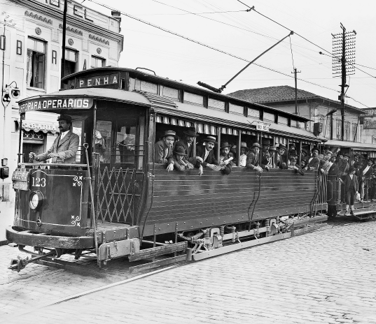 Imagem: Fotografia em preto e branco. Destaque de um bonde lotado. Na parte frontal, separado, está o condutor. Na segunda, os passageiros, em sua maioria homens, posam para foto nas janelas.  Fim da imagem.